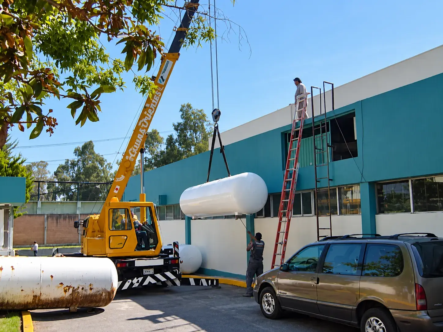 Grúa amarilla subiendo un tanque nuevo a la azotea de un edificio junto al tanque viejo oxidado
